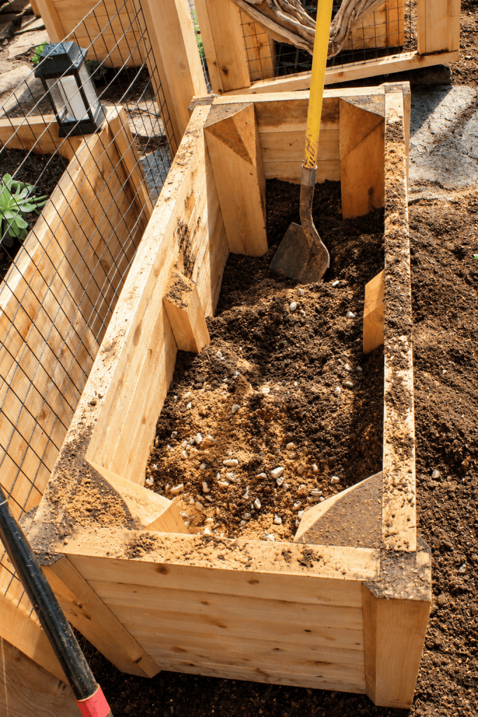 Close-up of a custom cedar raised garden bed being filled with soil and gravel, with a shovel inside and wire garden fencing surrounding the installation.