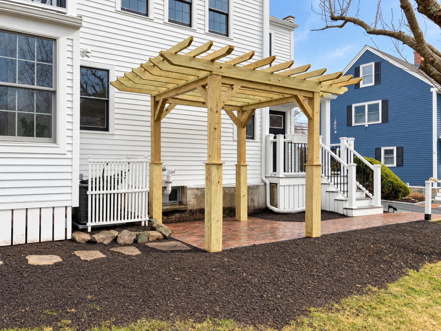Finished custom wood pergola over a brick patio beside a white home, surrounded by fresh mulch beds and landscaped outdoor space.