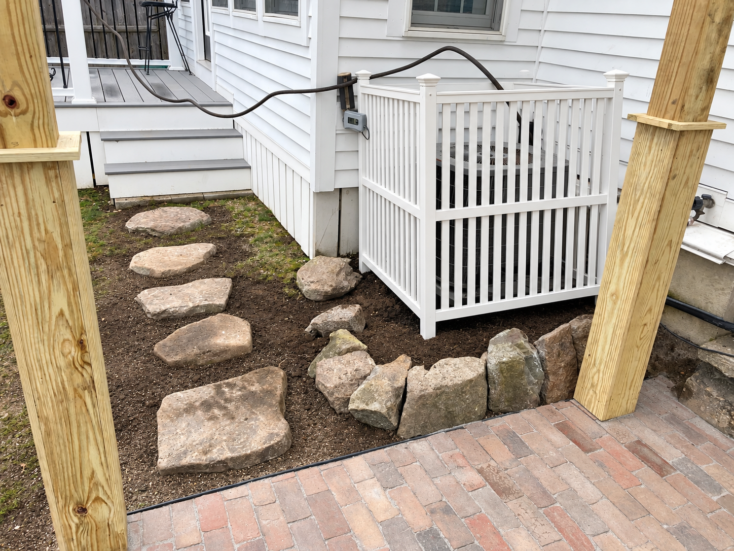 Brick patio beside a home with natural stone stepping stones, landscape edging, and a newly straightened white utility enclosure.
