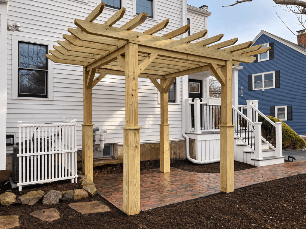 Newly built pressure-treated wood pergola over a brick patio beside a white residential home.