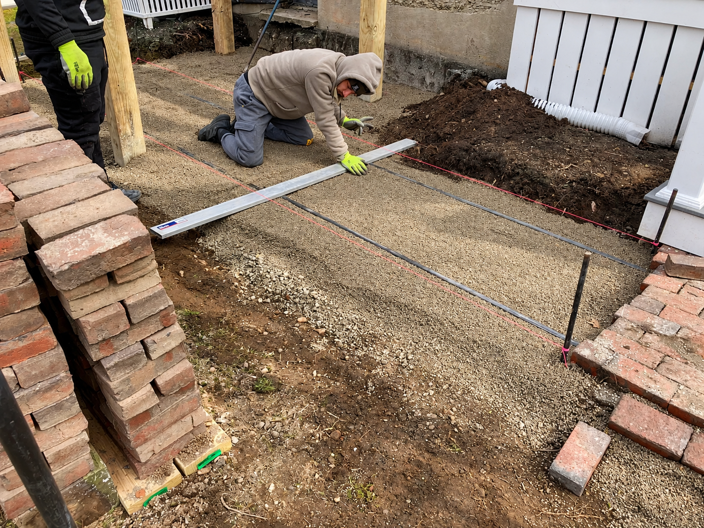 Landscaper leveling a compacted gravel base with guide strings beside stacked brick pavers during a residential patio installation.