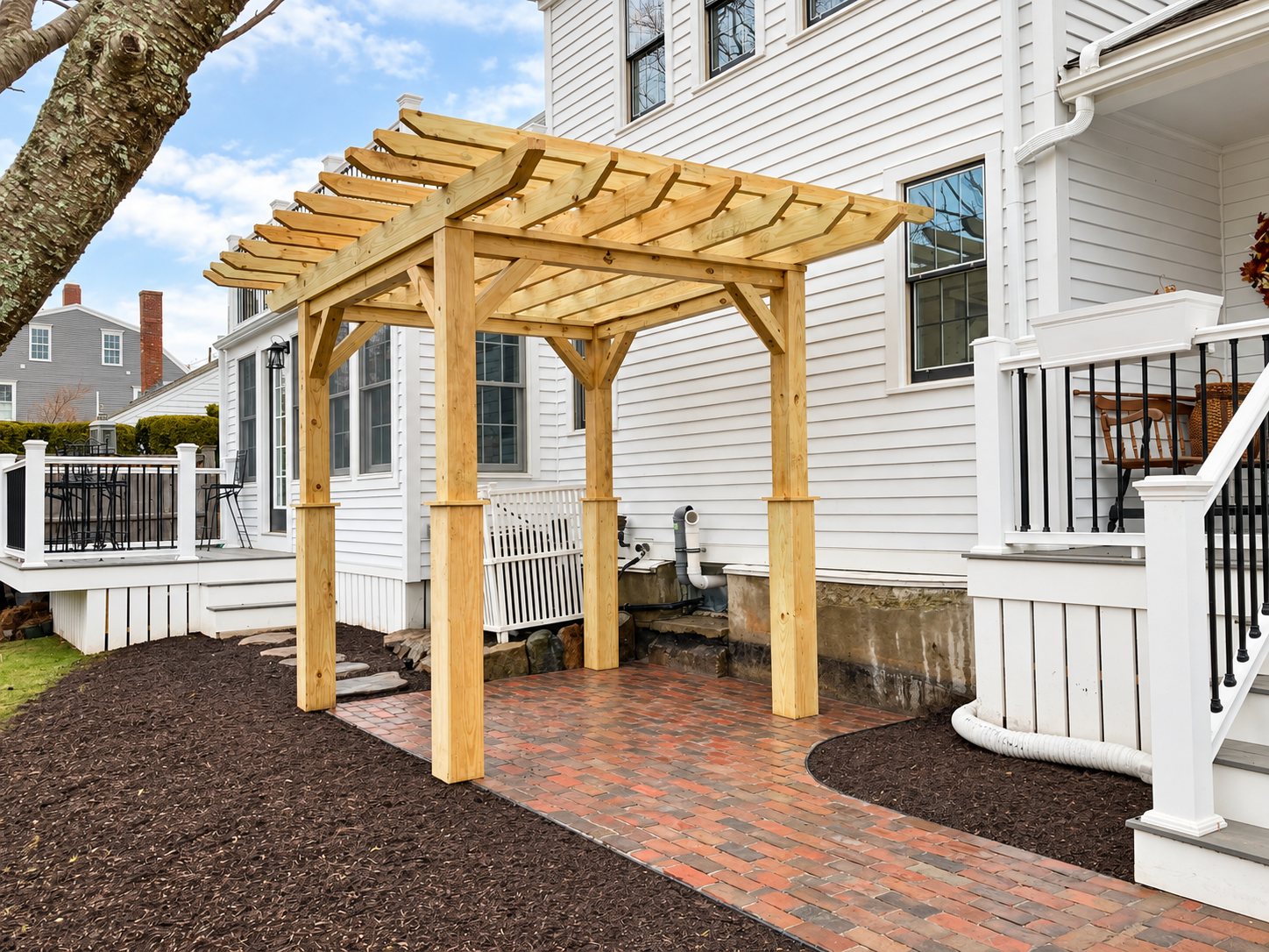 Finished custom wood pergola over a brick patio with fresh landscaping beside a white New England home.