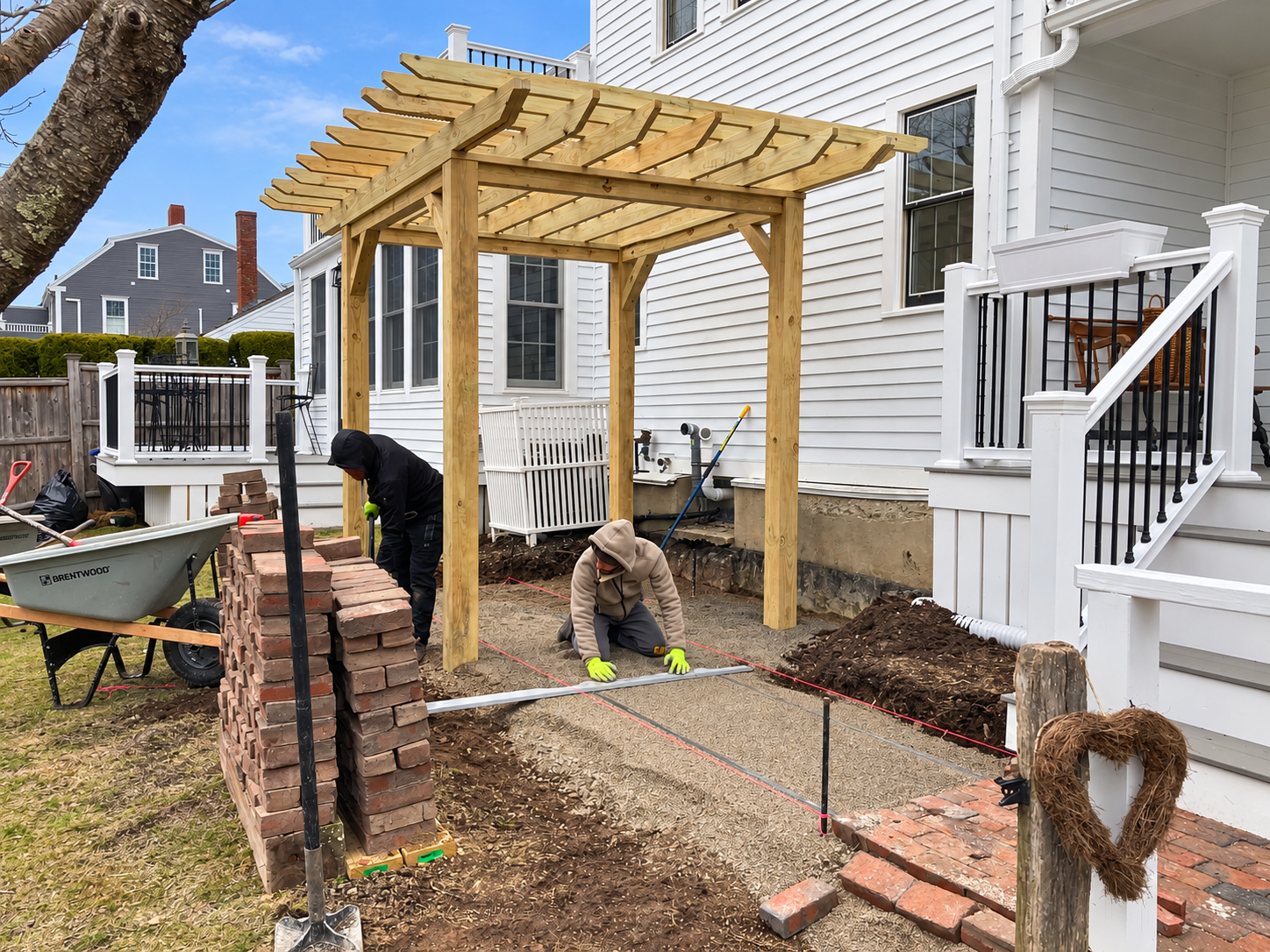 Landscaping crew preparing a brick patio base beneath a custom wood pergola beside a residential home.