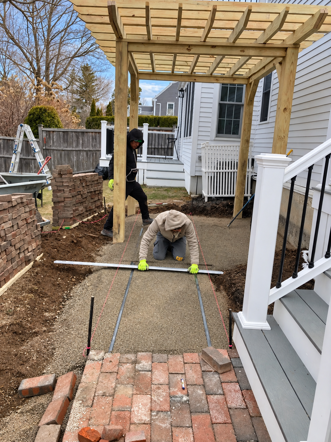 Landscaper leveling a compacted gravel base with guide strings beside stacked brick pavers during a residential patio installation.