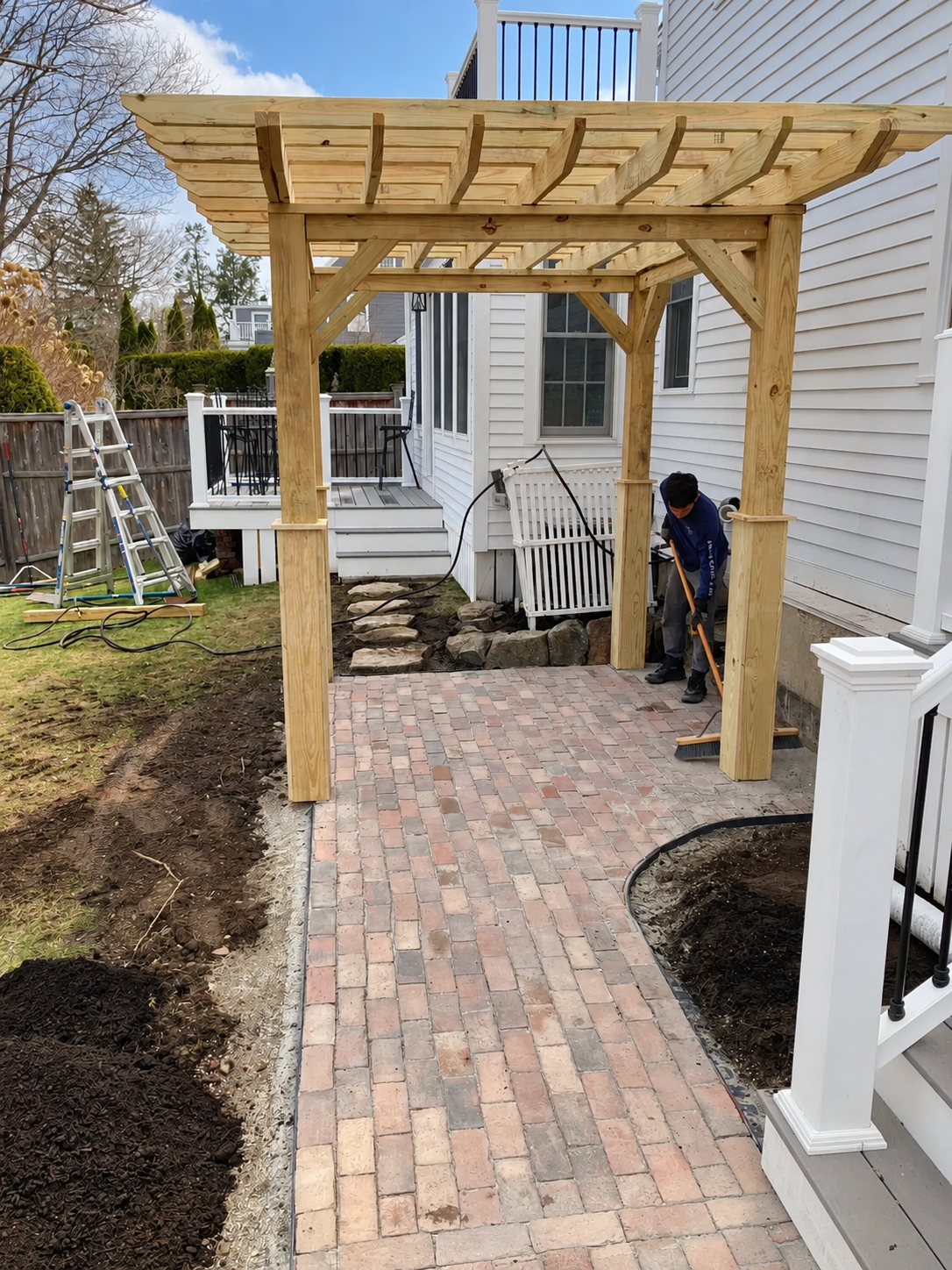 Landscaper sweeping a newly installed brick patio beneath a custom wood pergola beside a white home.