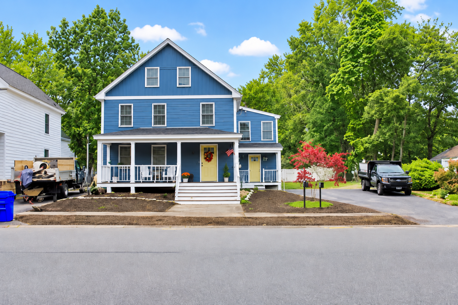 Blue two-story home with a white front porch, newly landscaped front yard, paved driveway, mature trees, and a small ornamental tree.