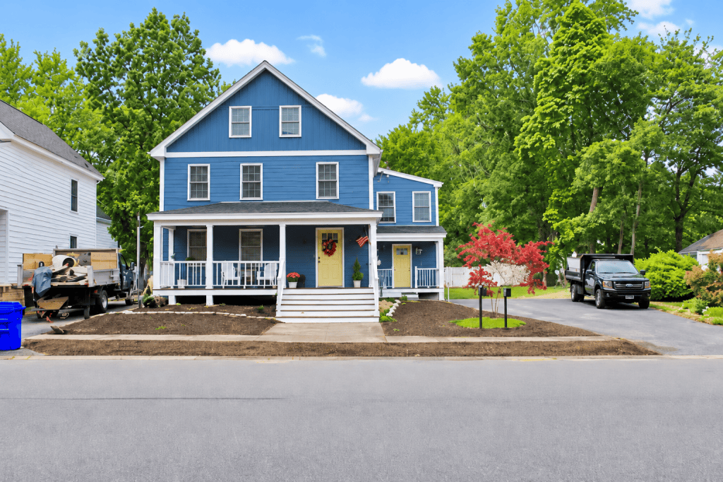 Blue two-story home with a white front porch, newly landscaped front yard, paved driveway, mature trees, and a small ornamental tree.