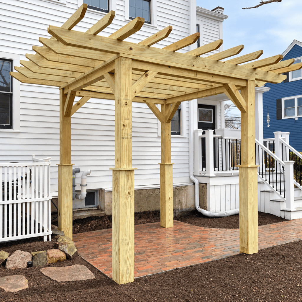Custom-built wood pergola installed over a brick patio beside a white home, with landscaped garden beds and porch steps nearby.