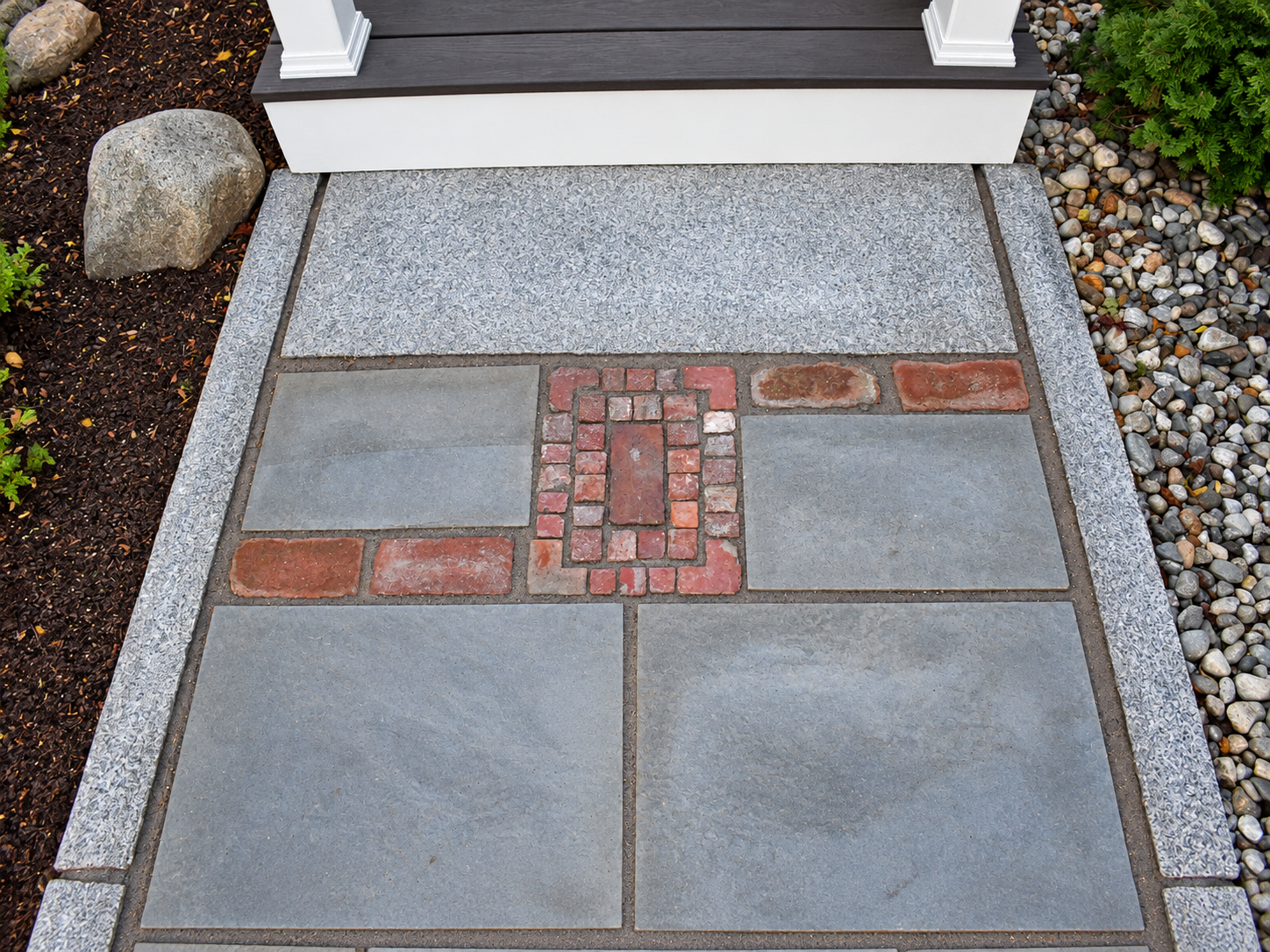 Close-up of a custom bluestone walkway with decorative red brick inlays and granite edging surrounded by garden beds.