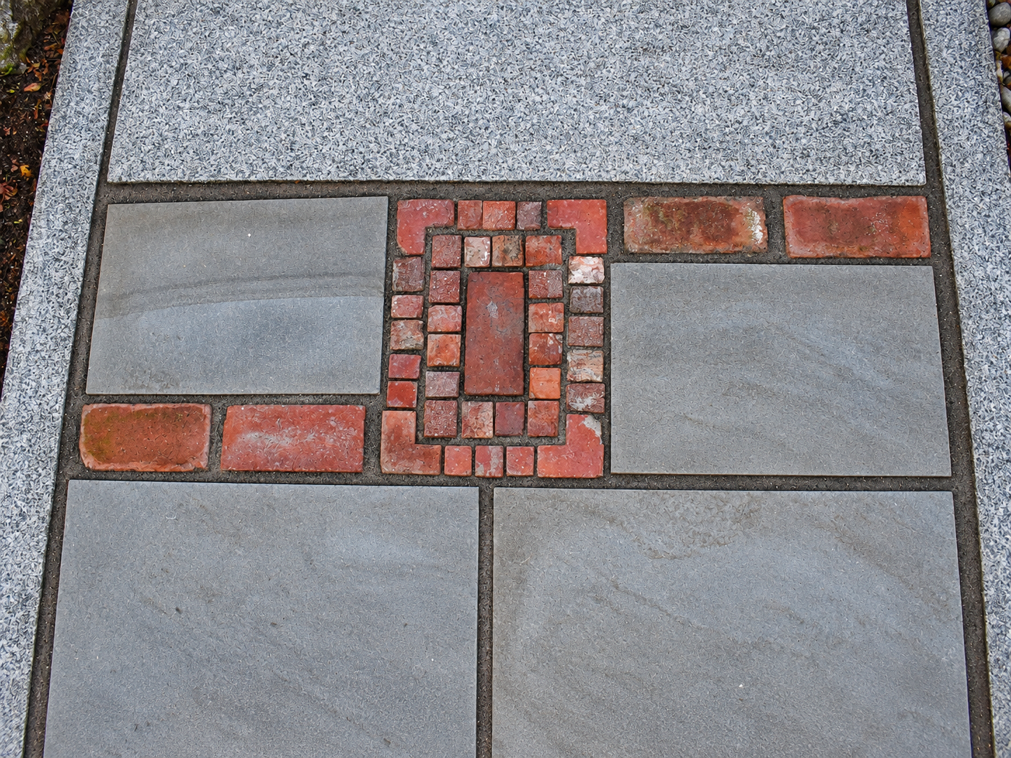 Close-up of a custom bluestone walkway with decorative red brick inlays and granite edging surrounded by garden beds.