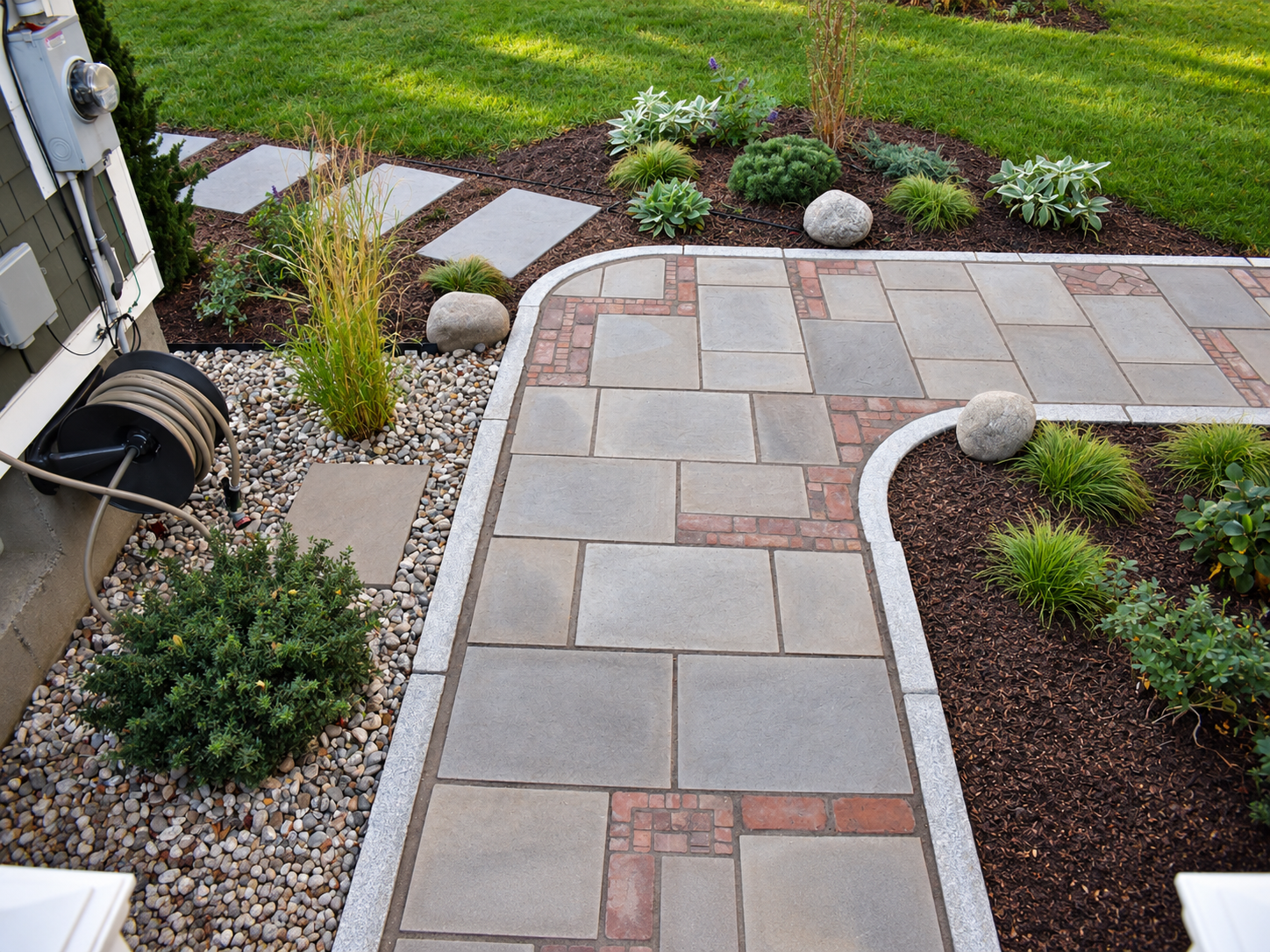 Close-up of a custom bluestone walkway with decorative red brick inlays and granite edging surrounded by garden beds.